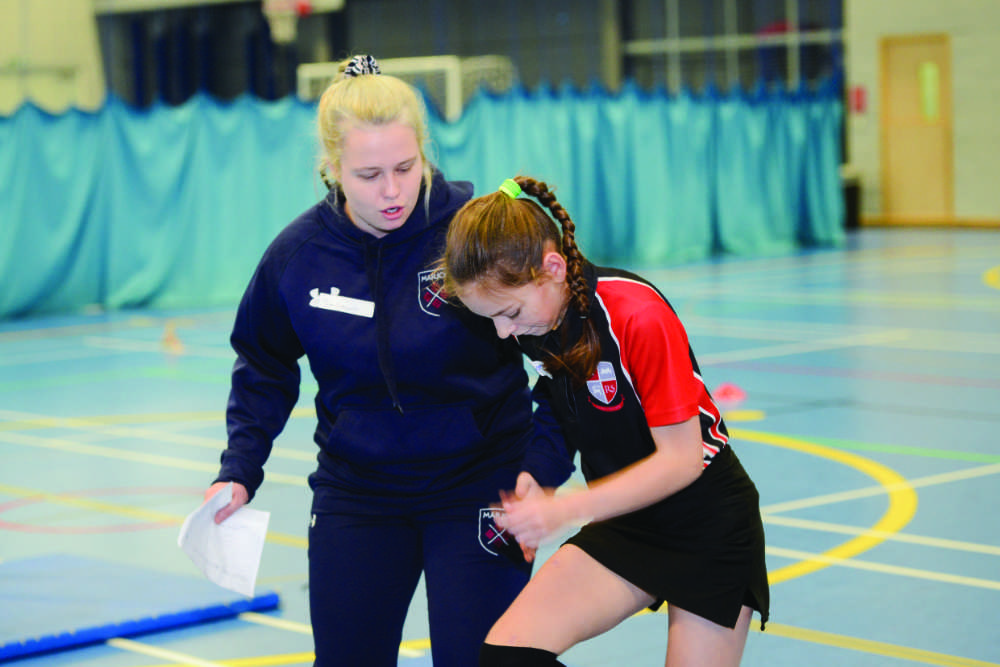 A student in a tracksuit coaching a younger student on a netball court