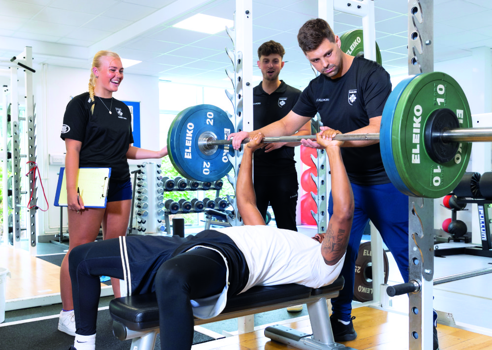 A group of Marjon sports students supervising someone benching weights in the Marjon Sport and Leisure Centre