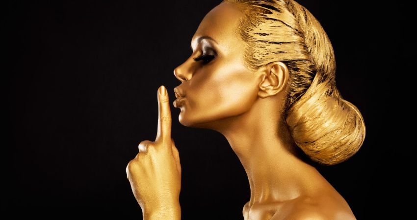 A woman painted gold who has her finger to her lips in a shushing gesture