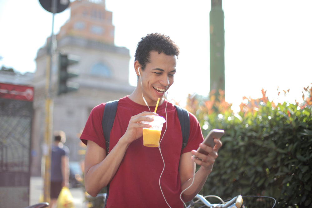 A mixed-race young person in a red t-shirt drinking an orange juice and looking at his phone