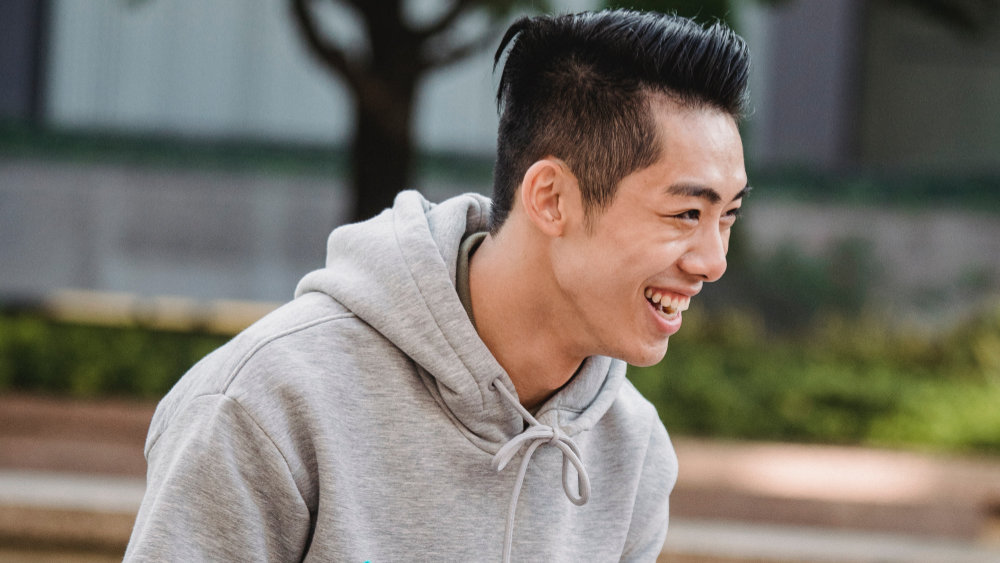 A young man in a grey hoodie smiles at something off-camera