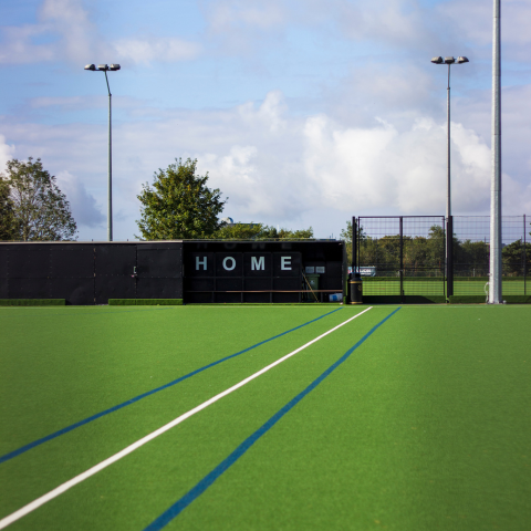 A green astroturf, bathed in sunshine with a black 'home' dug out in the distance
