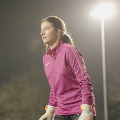 A Marjon student in a pink top and white football gloves standing on a floodlit pitch
