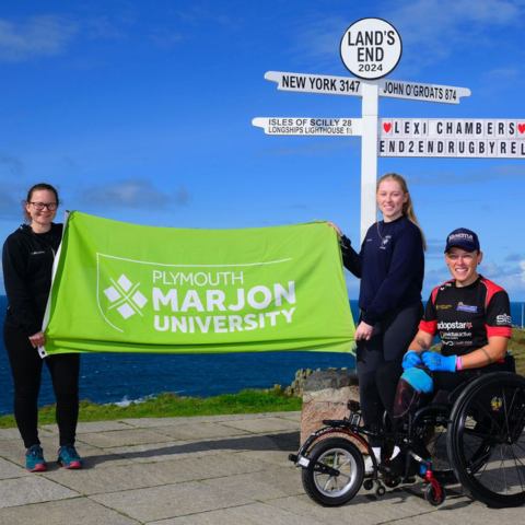 From left to right: Vicki Evans, Marjon lecturer, Libby Hill, Marjon student, and veteran Lexi Chambers holding a green flag with the Marjon logo on it in white, in front of the Land's End signpost.